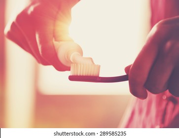 Hands holding a toothbrush and placing toothpaste on it in morning sunrise