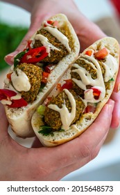 Hands Holding Two Halves Of Pita Bread Sandwich With Fresh Authentic Falafel Balls Inside, Chopped Salad And A Drizzle Of Tahini Sauce On Top. Selective Focus Of Chickpea Falafels In A Fluffy Pita.