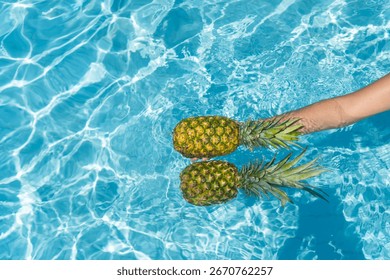 Hands holding two pineapples floating in a refreshing blue swimming pool on a sunny summer day