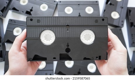 Hands Holding A VHS Tape Back Side With Scattered Tapes In The Background.