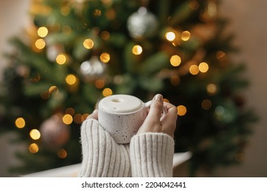 Hands holding warm cup of coffee on background of christmas tree with lights. Cozy home, atmospheric winter hygge. Woman hands in warm sweater holding  stylish mug at illumination bokeh - Powered by Shutterstock