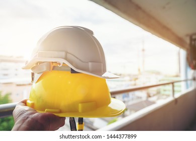 Hands holding a white and yellow helmet with a building under construction are blurred backgrounds. - Shutterstock ID 1444377899