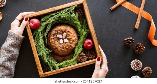 Hands holding a wooden box with a festive cake surrounded by greenery, apples, and pinecones. Cozy, rustic holiday cake setting with natural decorations. Woman unboxing Christmas pudding gift. - Powered by Shutterstock