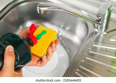 Hands holding a yellow scouring pad while pouring a splash of green dishwashing soap. Close up shot of a man's hands performing housekeeping duties. Concept of cleanliness