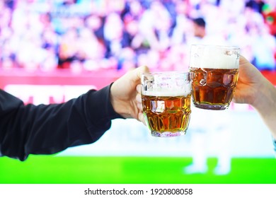 Hands Horizontal Holding Lager Beer Glass And Clinking On Background Of Football Game. Sport Fans Cheer Up. Friends Leisure Lifestyle Concept.