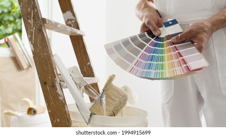 hands of house painter man decorator choose the color using the sample swatch, work of the house to renovate, a wooden ladder with paint brushes and a bucket as a background, close up
