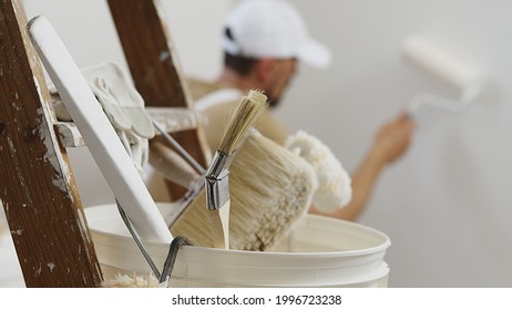 Handarbeit von Hausmalern, die von zu Hause zu renovieren ist, mit Rollerfarbe, weißer Eimer auf hölzerner Leiter mit Farbbürsten als Hintergrund, Nahaufnahme
