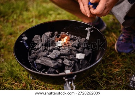 Hands ignite a starter cube among charcoal pieces in a small kettle grill outdoors on grass, with flame and light smoke for camping barbecue and cookout prep.