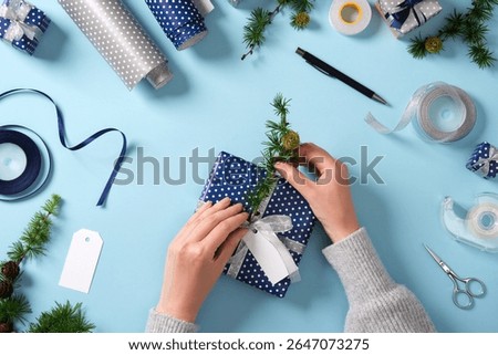 Hands insert a larch twig into a gift. Around it are ribbon, scissors, tape, and small wrapped gifts. Preparing for the holidays.