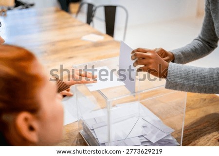 Hands inserting ballots into a clear ballot box on a table, illustrating citizens participating in elections, civic duty, secrecy and democratic choice for government and community