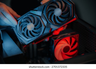 Hands installing graphics card inside desktop, focused DIY technician aligning high-performance GPU above glowing red case fan, close-up low-light scene showing