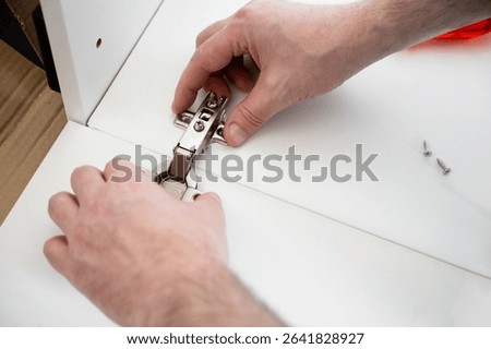 Hands installing a metal hinge on a white cabinet door with two screws nearby