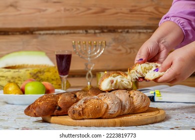 The Hands Of A Jewish Woman Break Challah After Kiddush With A Blessing Next To A Glass Of Wine And A Menorah. Horizontal Photo