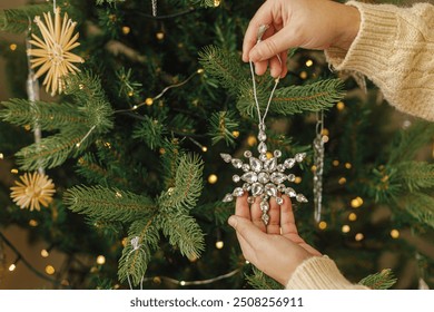 Hands in knitted sweater hanging stylish Christmas ornament on christmas tree with golden lights. Decorating christmas tree with vintage glass snowflake - Powered by Shutterstock