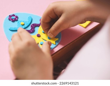 The hands of a little caucasian girl glue with her hands a blue felt sticker circle on a yellow bow easter egg with a lilac zigzag and a hat, sitting at a children's table with a pink background and d - Powered by Shutterstock