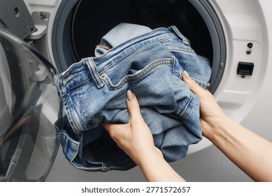 Hands loading blue jeans into washing machine drum at home laundry room. Household cleaning concept. Denim clothing wash scene. Domestic appliance closeup view. Laundry routine backdrop.