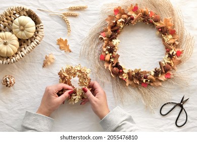 Hands Making Dried Floral Wreath From Dry Pampas Grass And Autumn Leaves. Flat Lay On Off White Textile Table Cloth. Scissors, Decorative Pumpkins And Dry Oak Leaves On The Table. DIY Fall Decorations