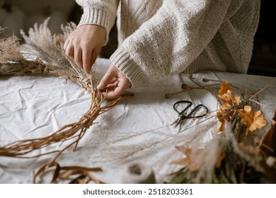 Hands making stylish autumn wreath. Dried grass, herbs, leaves, ribbon and scissors on rustic table. Details of fall decor and arrangement in home. Thanksgiving and Halloween - Powered by Shutterstock