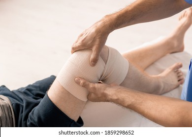 Hands Of Male Clinician Wrapping Knee Of Disable Patient With Flexible Bandage During Medical Procedure Before Rehabilitation Physical Training