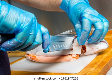 The Hands Of A Male Cook In Hygienic Gloves Cut Off The Head Of A Salmon With A Knife. Cutting Fresh Fish In The Kitchen On A Cutting Board.