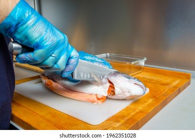 The Hands Of A Male Cook In Hygienic Gloves Cut Off The Head Of A Salmon With A Knife. Cutting Fresh Fish In The Kitchen On A Cutting Board.