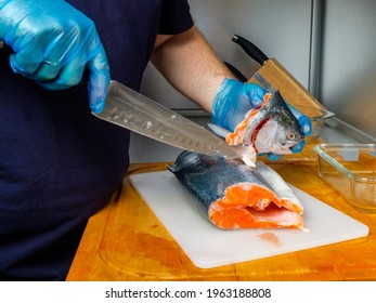 The Hands Of A Male Cook In Hygienic Gloves Cut Off The Head Of A Salmon With A Knife. Cutting Fresh Fish In The Kitchen On A Cutting Board.