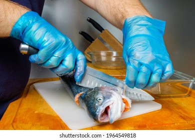 The Hands Of A Male Cook In Hygienic Gloves Cut Off The Head Of A Salmon With A Knife. Cutting Fresh Fish In The Kitchen On A Cutting Board.