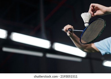 Hands of male player holding racket and hitting shuttlecock on dark badminton indoor court