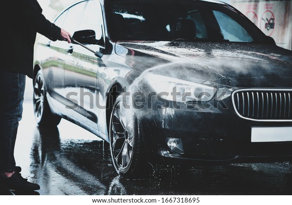 Hands\
of a male washing black car outdoors in a carwash station, using\
water jet with soap and high pressured water\
stream