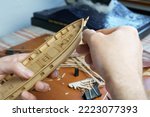 Hands of man cutting out details for ship model with clerical knife from plywood. Process of building toy ship, hobby and handicraft. Table with various materials, parts and devices for work