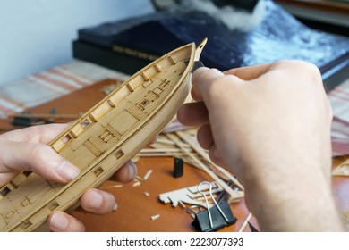 Hands of man cutting out details for ship model with clerical knife from plywood. Process of building toy ship, hobby and handicraft. Table with various materials, parts and devices for work - Shutterstock ID 2223077393