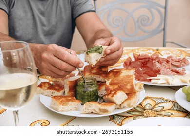 Hands of a man dipping bread into pesto sauce while enjoying a gourmet meal with assorted meats and cheeses on a beautifully decorated table