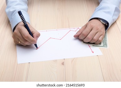 The Hands Of A Man Dressed On Blue Office T-shirt With Classic Wrist Watch And Golden Ring On His Hands, Draws A Chart Of Rising Stock Prices Using Red Pencil On The Table