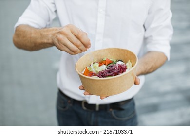 Hands Of Man, Eating Healthy Lunch Outdoor. Unrecognizable Male Torso In White Shirt, Hand With Fork, With Vegetable Salad In Paper Bowl, On Background Of Street, Diet Food Concept.