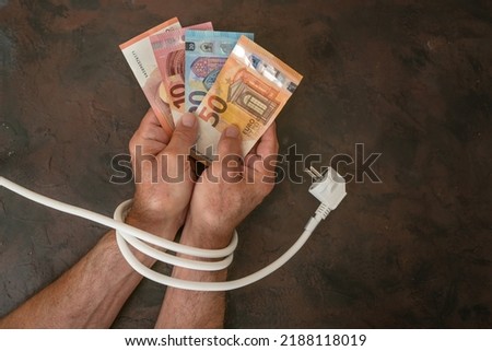 Hands of a man with euro banknotes and are tied up with an electric power cable, concept for rising electricity costs and saving energy, dark background, copy space, top view, selected focus