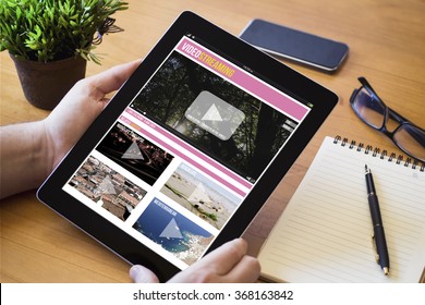 Hands Of A Man Holding A Video Streaming Device Over A Wooden Workspace Table. All Screen Graphics Are Made Up.