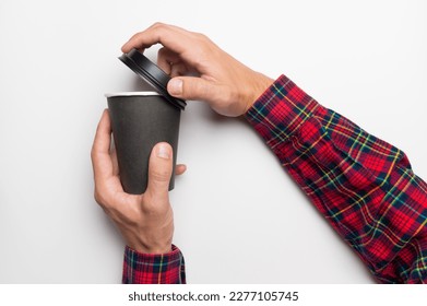 Hands of a man in a red plaid shirt with a paper black cup of coffee or tea on a white background, top view. The hand removes the plastic lid from the hot drink so as not to get burned - Powered by Shutterstock