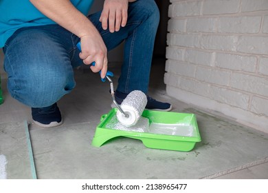 The Hands Of A Man Squatting Are Picking Up White Paint On A Construction Roller For Priming The Walls In The Room.