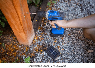 Hands of a man using a cordless drill to install a solar outdoor light on a wooden post. Concept of DIY, renewable energy, and eco-friendly garden lighting.