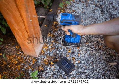 Hands of a man using a cordless drill to install a solar outdoor light on a wooden post. Concept of DIY, renewable energy, and eco-friendly garden lighting.