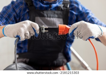 Hands of a man wearing work gloves hold an extension cord and cable connection of two plugs plugging the equipment into electricity.