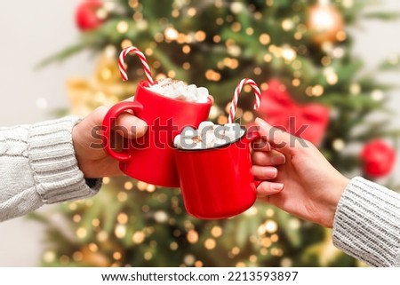 the hands of a man and a woman hold two red mugs with a hot drink and marshmallows on the background of the Christmas tree