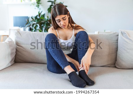 Hands massaging swollen foot while sitting on sofa during the day at home. Photo of Young Caucasian woman suffering from pain in leg. Woman massaging her legs after all day at work in office