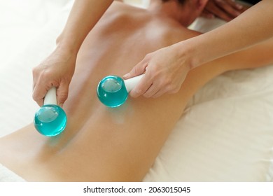 Hands of masseuse pressing two cold glass balls against female back during procedure of cooling massage