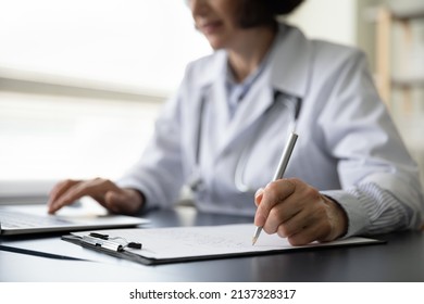 Hands of mature doctor woman filling paper medical records. Practitioner in white coat doing paperwork at workplace with laptop, writing notes, preparing documents, reports, prescription. Close up