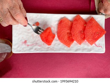 Hands Of Mature Woman Eating Watermelon In Triangular Pieces On A White Plate With Fork On A Pink Background Of A Paper Tablecloth Above A Table Of A Bar On The Terrace In Summer