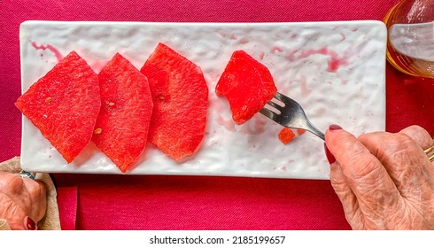 Hands Of Mature Woman Eating Watermelon In Triangular Pieces On A White Plate With Fork On A Pink Background Of A Paper Tablecloth Above A Table Of A Bar On The Terrace In Summer