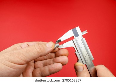 Hands measuring small screw using a metal caliper against a red background. Right hand adjusts the caliper to determine the screw's diameter. Precision instrument used in workshops engineering fields 
