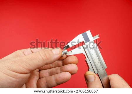 Hands measuring small screw using a metal caliper against a red background. Right hand adjusts the caliper to determine the screw's diameter. Precision instrument used in workshops engineering fields 