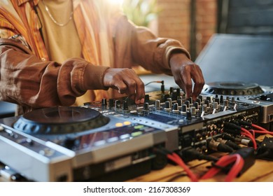 Hands of modern black man regulating sounds on dj set while standing by table in studio or loft apartment and turning mixers